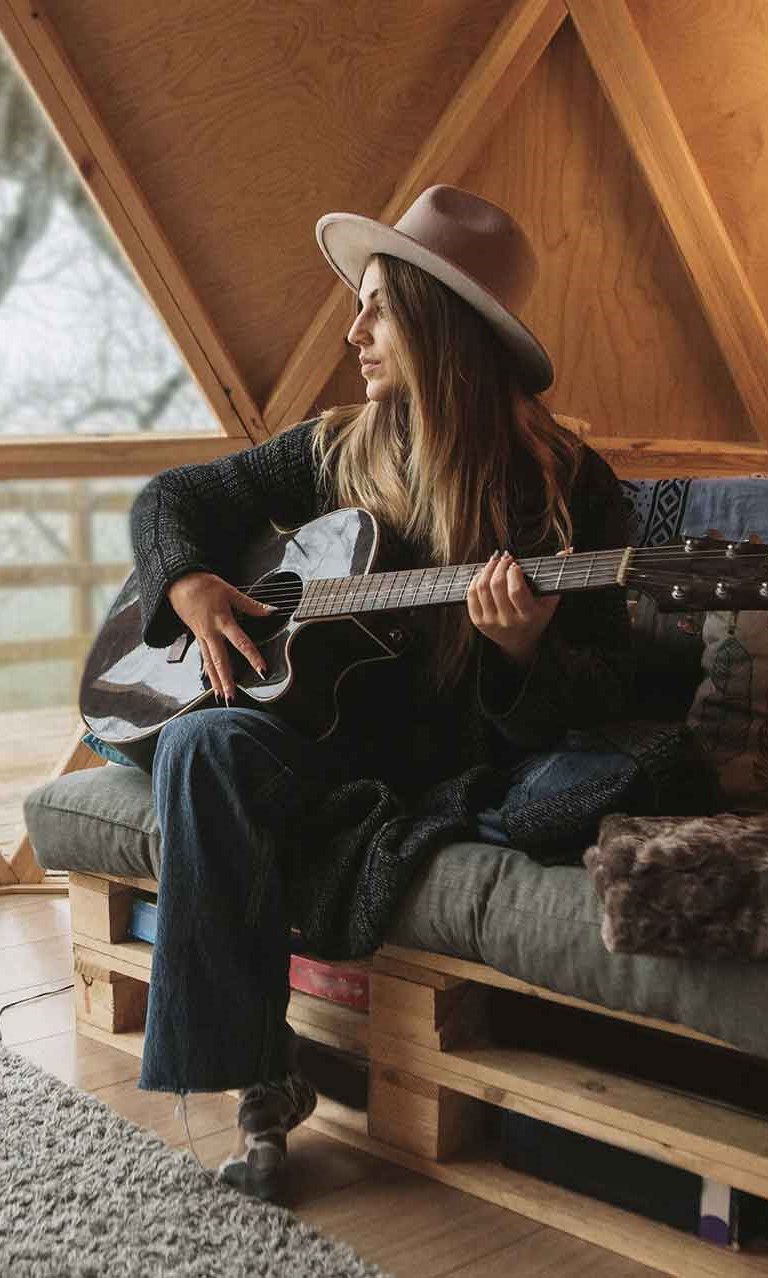 vrouw op een zelfgemaakte sofa die gitaar speelt en uitkijkt naar buiten in een alternatieve woning (tiny house)
